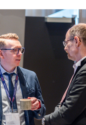 Two men in suits are having a conversation at an event. One of them is holding a coffee mug and wearing a name badge that reads "Tom Lehane, Epical Sweden AB." Both are wearing event lanyards. Other attendees are visible blurred in the foreground.